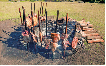 Imagem: Fotografia. Local com grama de cor verde. Ao centro, solo de cor marrom-escuro, com hastes de madeiras na vertical e nelas, pedaços de carnes de cor marrom fincados e linguiças. Ao fundo, toras de madeiras em marrom. Fim da imagem.