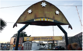 Imagem: Fotografia. Entrada de local com portões e grades, com a parte superior com faixa em marrom-claro, com texto: BEM-VINDOS. Na parte superior, em cima de duas hastes de cor preto, com chapéu de cangaceiro de cor marrom com a parte superior arredondada, com três estrelas em branco. Na parte superior, céu claro em azul-claro.  Fim da imagem.