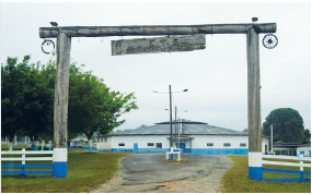 Imagem: Fotografia. À frente, entrada de local com hastes na vertical e na parte superior na horizontal, em madeira de cor marrom. Ao centro, no alto, placa em marrom onde está escrito C. T. G. À esquerda e à direita, cercado em branco e azul. Mais ao fundo, um galpão de paredes de cor branca e parte inferior em azul, com telhado cinza na parte superior. À esquerda e à direita, árvores de folhas verdes. No alto, céu claro.  Fim da imagem.