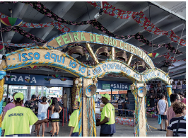 Imagem: Fotografia. Ao centro, placa com a inscrição: Festa de São Cristovão. E faixas coloridas e estampadas. Abaixo, diversas pessoas. Fim da imagem.