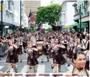 Imagem: Fotografia. Rua cinza, com centenas de pessoas desfilando, à frente, mulheres usando vestidos de cor preta com detalhes em branco, com o braço direito para cima. Elas têm pele clara, a maioria de cabelos escuros. Mais ao fundo, homens também usam camiseta preta e branca. Mais ao fundo, à direita, árvore de folhas verdes e prédios à esquerda e à direita. Sobre alguns, sacadas com pessoas observando o desfile.  Fim da imagem.