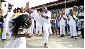 Imagem: Fotografia 3. À esquerda, duas crianças negras de cabelos pretos com pequenas presilhas, usando camiseta e calça de cor branca, com os pés descalças. À esquerda, menina agachada e à frente, menina com a perna esquerda perpassando sobre o corpo da outra. Mais ao fundo, outros meninos e meninas, alguns vestidos com camiseta e calça em branco. Mais ao fundo, vista parcial de local com telhado.  Fim da imagem.