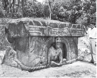 Imagem: Fotografia em preto e branco. À esquerda, altar de pedra na horizontal de cor cinza, com silhueta de uma pessoa na parte inferior, sentado com as pernas cruzadas. No alto, parte na horizontal. À direita, homem em pé de cabelos escuros, com camiseta branca, calça clara e mão esquerda sobre a pedra. Em segundo plano, local com árvores de folhas.  Fim da imagem.