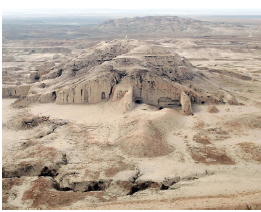 Imagem: Fotografia. Vista do alto de local com solo arenoso de cor bege-claro e ao fundo, morros com pedras altas de cor bege, com aberturas na parte inferior, similares a portas.  Fim da imagem.