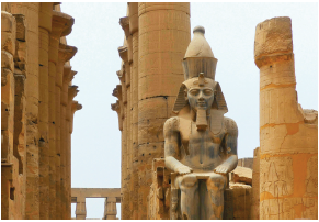 Imagem: Fotografia. Vista geral de local com vigas de cor bege na vertical e à direita, uma estátua gigante de pessoa sentada, mãos sobre os joelhos, com chapéu sobre a cabeça. À direita, viga na vertical com parte superior quebrada. No alto, céu nublado cinza.  Fim da imagem.