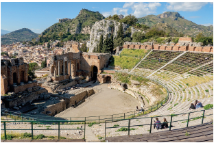 Imagem: Fotografia. Local com formato arredondado com as arquibancadas, com quatro pessoas sentadas. À frente e na parte do fundo, grades. Ao fundo, palco do anfiteatro, com pedras e paredes de cor marrom com partes quebradas. Em terceiro plano, casas e morros com vegetação verde, céu azul-claro e nuvens brancas.  Fim da imagem.