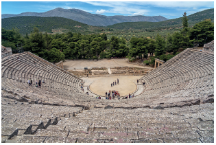 Imagem: Fotografia. Vista geral de arquibancadas de teatro com formato arredondado. Na parte inferior, palco com pessoas em pé. Em segundo plano, parte com solo em bege, com árvores de folhas verdes. Em segundo plano, morros e no alto, céu em azul-claro e nuvens brancas.  Fim da imagem.