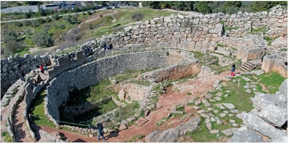 Imagem: Fotografia. Vista do alto de local com ruinas, de cor cinza, de vegetação rasteira verde, com paredão contornando na horizontal. Ao fundo, pedaços de pedras cinzas. Mais ao fundo, à esquerda, local com grama.  Fim da imagem.