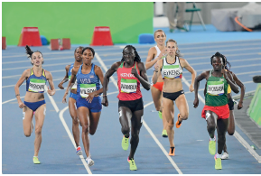 Imagem: Fotografia. Em uma pista de corrida de cor azul-claro, com linhas finas de cor branca. Correndo dezenas de mulheres com placa de numeração no peito, com short curto e blusa regata, de etnias diferentes: de pele clara, morena e negras.  Fim da imagem.