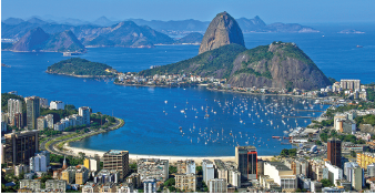 Imagem: Fotografia. Vista do alto de local com cidade vista parcialmente, prédios de tamanhos diversos, pequenos, médios e à frente, o mar de cor azul. Mais ao fundo, morros de cor cinza arredondados no mar. Em segundo plano, paisagem com outros morros e céu azul.  Fim da imagem.