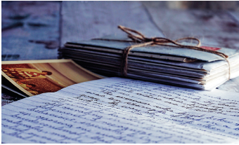 Imagem: Fotografia. À frente, uma folha branca com texto escrito em letra cursiva, com fotos à esquerda e mais ao fundo, envelopes amontoados e amarrados com um cordão marrom.  Fim da imagem.