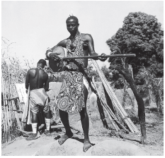 Imagem: Fotografia em preto e branco. À frente, um homem de pele negra, descalço com vestes compridas do pescoço até os joelhos, segurando nas mãos um instrumento nas mãos, com parte arredondada à esquerda e haste fina horizontal na mão direita. Mais ao fundo, à esquerda, um homem visto de costas, sem camiseta, com roupa clara, na cintura. À direita, tronco na vertical e mais ao fundo, árvores de folhas na copa no alto.  Fim da imagem.