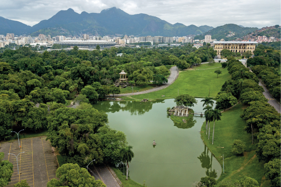 Imagem: Fotografia. Vista geral de local com rio ao centro de cor verde, com contornos com vegetação verde, na ponta da esquerda e à direita, árvores. Nas extremidades, ruas de cor cinza. Em segundo plano, mais vegetação e prédio de paredes de cor bege. Em terceiro plano, prédios da cidade, morros e no céu, nuvens de cor cinza. As plantas nas áreas verdes contribuem para melhorar a qualidade do ar, diminuindo a quantidade de gás carbônico na atmosfera. Ao realizar a fotossíntese, as plantas absorvem o gás carbônico e liberam o gás oxigênio. O solo coberto por gramados favorece a infiltração da água da chuva, evitando alagamentos, enquanto as raízes das plantas protegem o solo da erosão. Fim da imagem.