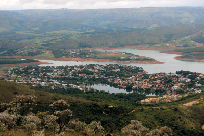 Imagem: Fotografia. Vista do alto de local com morros ao redor, com vegetação, árvores de folhas verdes e ao centro, local com rio e perto dele, casas e árvores. Mais ao fundo, rios e morros. No alto, céu visto parcialmente com nuvens cinzas.  Fim da imagem.