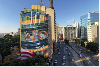 Imagem: Fotografia. Vista do alto de cidade, com prédios altos à esquerda, um deles, com ilustração colorida de rosto de uma pessoa, estampado e à direita, prédio espelhado. Ao centro, rua de cor cinza, com muitos carros passando. No alto, céu de cor azul-claro, com pequenas nuvens à direita de cor branca.  Fim da imagem.