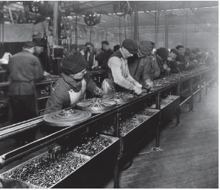 Imagem: Fotografia em preto e branco. Dezenas de homens de frente para uma base com rodas de ferro de automóveis. Na parte inferior da base, dezenas de peças pequenas de ferro, similares às roscas. Ao fundo, outros homens trabalhando, a maioria usa roupa antiga, com boina na cabeça.  Fim da imagem.