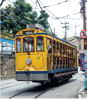 Imagem: Fotografia. Em uma rua de cor cinza, com trilho de ferro na rua, por onde passa um bondinho de cor amarela, com cabos finos, na vertical passando em fios pendurados na horizontal. Dentro do bonde, pessoas sentadas e uma pessoa em pé dentro. Em segundo plano, cidade, carros e árvore de folhas verdes. Na ponta da direita, poste com placa de proibido estacionar e emaranhados de fios pretos de energia. O céu tem cor cinza-claro.  Fim da imagem.