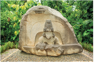 Imagem: Fotografia. Uma pedra grande de cor cinza, arredondada. À frente, escultura com a cabeça arredondada, chapéu sobre cabeça. Em segundo plano, local com densa vegetação.  Fim da imagem.