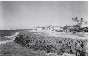 Imagem: Fotografia em preto e branco. À esquerda, praia com ondas e à direita, local com pedras e mais ao fundo, casas de paredes brancas, com telhado e árvores grandes com folhas na parte superior. Fim da imagem.