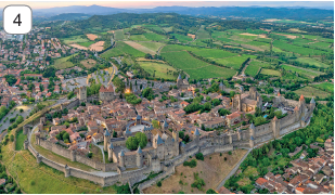 Imagem: Fotografia 4. Vista do alto de local com um muro grande de cor cinza, contornando parte com muitas casas dentro, antigas de telhados em marrom e um castelo à esquerda com torres na vertical, com ponta superior em azul-claro. Dentro, entre algumas casas, árvores com folhas verdes. Ao redor, fora da área contornada com muro, com vegetação verde à direita e à esquerda, outras moradias entre árvores. Em segundo plano, morros e vegetações. Fim da imagem.