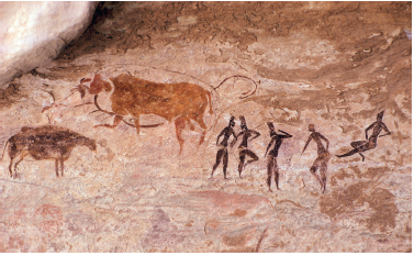 Imagem: Fotografia. Sobre uma rocha de cor marrom, com pinturas rupestres de cor marrom. À esquerda, dois animais rupestres, um similar a um animal bovino. À direita, cinco silhuetas de cor marrom-escuro, quatro em pé e outra à direita, sentado.  Fim da imagem.