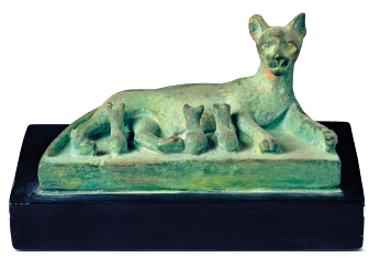 Imagem: Fotografia. Uma estátua de cor verde-claro sobre base na horizontal de uma gata com orelhas pequenas, com o corpo deitado e de frente para ela, filhotes amamentando.  Fim da imagem.