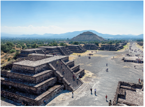 Imagem: Fotografia. Vista do alto de local com solo de cor cinza e à esquerda, ruinas em cinza com o formato retangular com degraus diferentes e ao centro, uma escadaria grande na vertical, por onde passam pessoas. Ao fundo, outras ruinas similares, em segundo plano, morros e no alto, céu em azul-claro.  Fim da imagem.