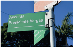 Imagem: Fotografia. Um poste na vertical de cor cinza, com duas placas verdes. À frente, placa com letras brancas: Avenida Presidente Vargas. Ao fundo, árvore com folhas verdes e céu em azul-claro.  Fim da imagem.