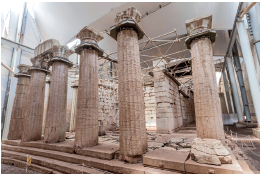 Imagem: Fotografia. Vista de baixo para cima, de local em ruinas de cor bege com base com degraus e colunas na vertical, sem teto e ao fundo, hastes de ferro e paredes em ruinas.  Fim da imagem.