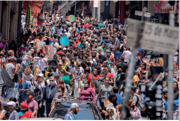 Imagem: Fotografia. Vista do alto de dezenas de pessoas aglomeradas em uma rua. Algumas usam máscara para tampar nariz e boca, homens e mulheres. Na ponta da direita: poste cinza na vertical, com uma placa da mesma cor escrita: 25 de março.  Fim da imagem.