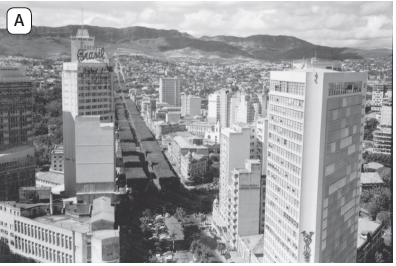 Imagem: Fotografia em preto e branco A. Vista do alto de local com rua ao centro grande com algumas árvores, com prédios à esquerda e à direita, de tamanho médio, pequeno e outros altos. Mais ao fundo, em segundo plano, morros com vegetação. No alto, céu com nuvens brancas.  Fim da imagem.