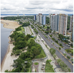 Imagem: Fotografia. Vista do alto de local com vegetação esquerda, árvores à esquerda, perto de um rio de água de cor escura. À direita, ruas de cor cinza por onde passam automóveis. Mais à direita, locais com vegetação, árvores e prédios modernos altos. No alto, céu nublado com nuvens de cor cinza.  Fim da imagem.