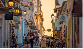 Imagem: Fotografia. Vista geral de local com uma rua ao centro por onde passam pessoas e à direita, outras pessoas sentadas. A rua não é muito larda, com casas umas ao lado da outra, à esquerda e à direita, de paredes coloridas em azul, amarelo, rosa e outras cores. Perto das portas, luminárias acesas com luz de cor amarela. No alto, céu de cor amarelada. Fim da imagem.