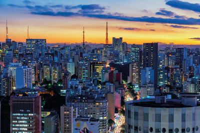 Imagem: Fotografia. Vista do alto de local com rua ao centro, vista parcialmente por onde passam carros e no entorno, prédios de tamanhos médios, altos e ao fundo, quadro torres de ferro na vertical. Alguns prédios, têm luzes acesas nas janelas. No alto, céu em tons de amarelo e à direita, nuvens escuras.  Fim da imagem.