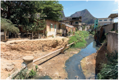 Imagem: Fotografia. Local com solo de cor bege à esquerda e à direita, rio de cor verde e cinza, com um cano fino na horizontal, com árvores à esquerda com folhas verdes. Mais ao fundo, casas aglomeradas entre si, inacabadas. Em segundo plano, morros grandes de cor verde. No alto, céu em azul-claro e à direita, vista parcial de cor branca.  Fim da imagem.