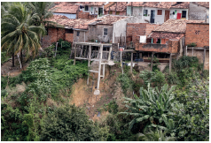 Imagem: Fotografia. Na parte inferior, morros de cor bege, com árvores e vegetação de cor verde. Na parte superior, casas inacabadas uma ao lado da outra, com paredes de cor cinza e marrom, algumas com telhados marrom e partes inferior, com hastes de madeira.  Fim da imagem.