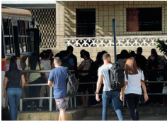 Imagem: Fotografia. À esquerda, vista parcial de um ônibus com janelas de vidro e à frente, porta aberta por onde entram dezenas de pessoas. Ao fundo, outras três pessoas perto de uma grade: homem de pele clara, camiseta azul e bermuda cinza, com mochila preta nas costas. Mais à direita, um homem de pele morena, cabelos pretos, com camiseta cinza, calça azul e mochila preta nas costas. Uma mulher de cabelos curtos castanhos, com camiseta de cor branca e calça preta. Em segundo plano, local com paredes de cor bege, com uma porta e uma janela.  Fim da imagem.