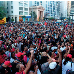 Imagem: Fotografia. Vista geral de local com pessoas aglomeradas na rua, muitas pessoas vestidas com camiseta e boné na cabeça de cor vermelha, a maioria são morenos de cabelos pretos, e tem a mão fechada para o alto. Em segundo plano, prédios de paredes de cor cinza e um estabelecimento de paredes beges com teto triangular e uma cruz no alto.  Fim da imagem.