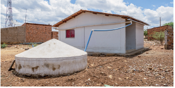 Imagem: Fotografia. Local com solo de cor marrom-claro, com casa de paredes brancas, telhado triangular, com um cano de cor azul para uma cisterna, com o formato redondo e parte superior triangular. Mais ao fundo, à esquerda, casas de paredes de tijolo marrom. No alto, céu azul-claro com nuvens brancas.  Fim da imagem.