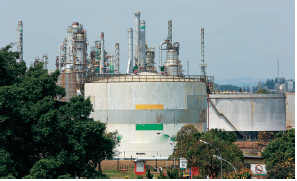 Imagem: Fotografia. Vista geral de local com forma arredondada cinza, grande e mais ao fundo, estruturas na vertical e de formato tubular e ferros. À frente, local com solo marrom-claro, árvores pequenas de folhas em verde-claro e à esquerda, vista do alto de copa de árvore de folhas em verde-escuro. No alto, céu azul-claro sem nuvens.  Fim da imagem.