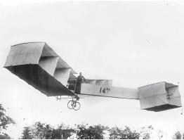 Imagem: Fotografia em preto e branco. Na parte inferior, vista do alto da copa de várias árvores e no alto, céu por onde sobrevoa um avião antigo, com estrutura na horizontal, com estrutura similar a tecidos finos claros, com blocos quadrado, haste fina à frente, ao centro, homem em pé o pilotando e na parte inferior, duas rodas pequenas. Na haste na horizontal está escrito: 14 bis.  Fim da imagem.