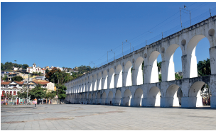 Imagem: Fotografia. Vista geral de local à esquerda, rua de cor cinza-claro e à direita, com muro branco com manchas pretas acima. Dentro, arcos arredondados por onde vê-se paisagem com céu azul claro e árvore de folhas verdes. Ao fundo, à esquerda, casas de paredes de cor branca, telhados marrons e  entre elas, árvores com folhas verdes.  Fim da imagem.