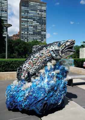 Imagem: Fotografia. Ao centro, uma escultura com peixe grande cor cinza, de lata, com o corpo dele cheio de tampas de garrafa plástica e abaixo, plástico de cor azul-claro, partes em branco de plástico e pedaços plásticos em azul. Em segundo plano, local com muro cinza, vegetação verde, árvore com folhas verdes e mais ao fundo, prédio visto parcialmente de cor cinza. À direita, céu azul-claro. Fim da imagem.