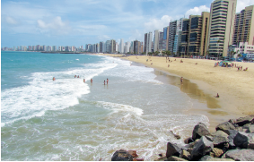 Imagem: Fotografia. À esquerda praia com água de cor azul, com ondas em branco, com pessoas e à direita, areia de cor bege-claro, pessoas na areia e na ponta da direita, pedra de cor cinza. Em segundo plano, prédios altos e médio da cidade com janelas, céu em azul-claro e nuvens brancas.  Fim da imagem.