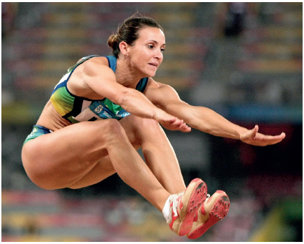 Imagem: Fotografia. Uma mulher usando uniforme. Ela está saltando com as pernas flexionadas e os braços e mãos estendidos para frente. Fim da imagem.