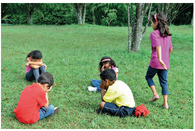 Imagem: Fotografia. Quatro crianças sentadas em roda fazendo um círculo. Eles estão com as mãos nos olhos. Ao lado, uma menina em pé andando ao redor deles. No fundo, a árvore e a grama. Fim da imagem.