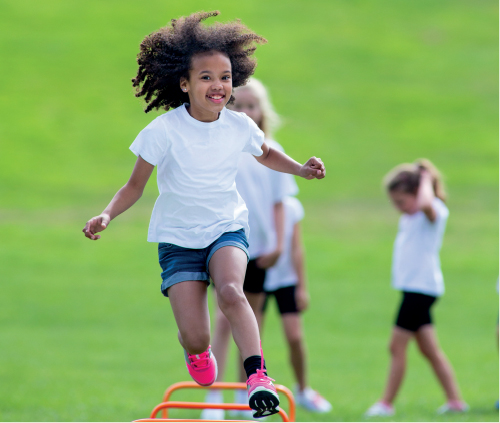 Imagem: Fotografia. Uma menina com cabelos crespos, uniformizada saltando sobre obstáculos. Atrás uma fila de crianças uniformizadas. Fim da imagem.