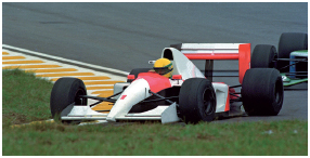 Imagem: Fotografia. Um piloto dentro de um carro de fórmula 1 (um carro baixo, com quatro rodas e uma asa atrás. Fim da imagem.