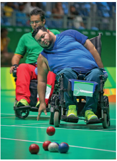 Imagem: Fotografia. Um homem usando camiseta azul, calça jeans, tênis. Ele está sentado em uma cadeira de roda com a mão na direção do chão e de uma bola. Na frente um conjunto de bolas coloridas. Fim da imagem.