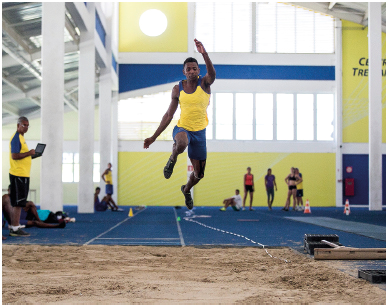 Imagem: Fotografia. Um homem uniformizado. Ele está saltando em uma pista azul. Na frente, um quadro com areia. Fim da imagem.